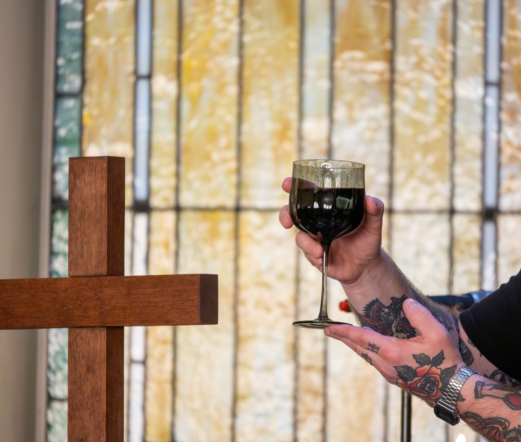 A hand raising the communion cup beside a wooden cross, stained glass behind.